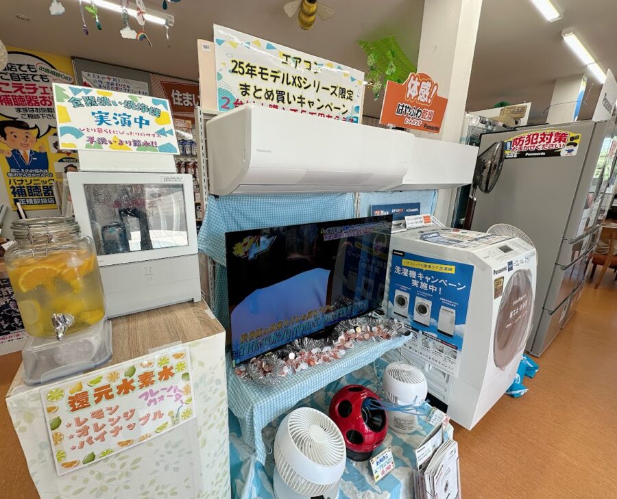 ﾊﾟﾅｿﾆｯｸ専門店の入り口を入ってすぐの展示風景。夏の式なので最新モデルのエアコンが目立つようbに展示され、その下に最新のテレビの展示、横にはドラム式洗濯乾燥機や冷蔵庫が並ぶ。左側には実演の一人用食洗機が稼働している。また、還元水素水で作ったフルーツのフレーバーウォーターが試飲できるようになっている。